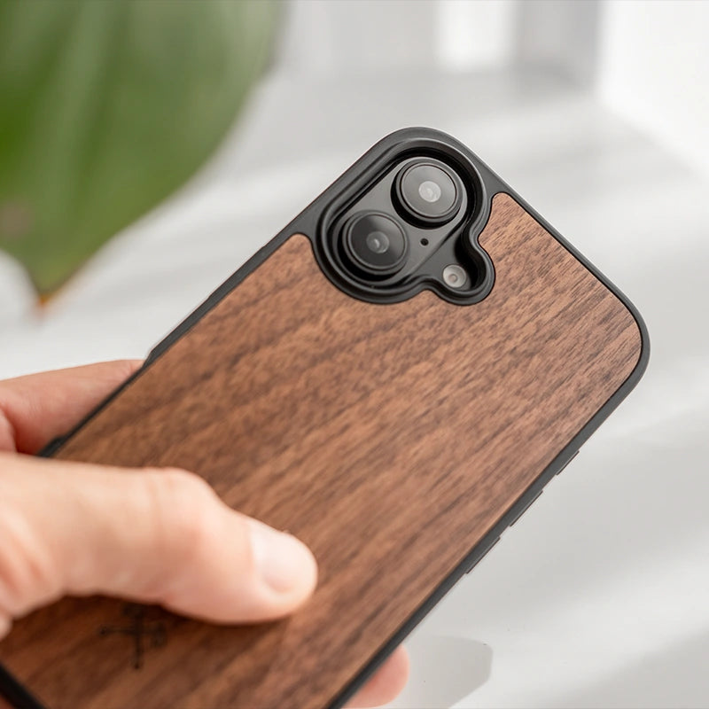 Gros plan sur une main tenant un iPhone 16 dans la finition « Bumper Case » en bois de noyer, sur un fond clair.