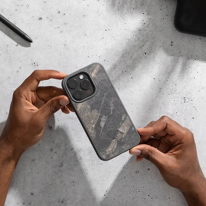 Gros plan sur une personne tenant un iPhone 16 Pro en mode « Bumper Case » avec une pierre, sur un fond clair.