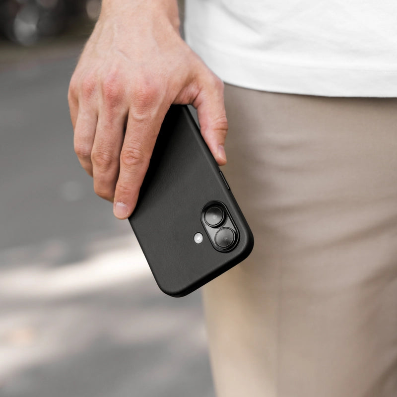Eine Hand hält ein schwarz iPhone 17 Air im schwarzen Leder-Case im Freien. Im unscharfen Hintergrund sind eine helle Hose und Straßenpflaster zu erkennen.