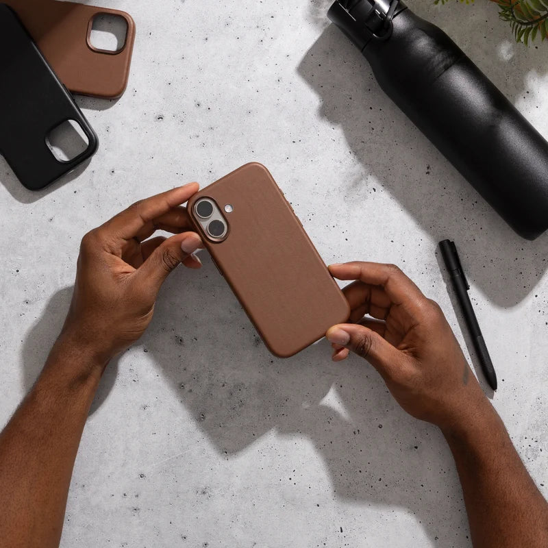 Zwei Hände halten ein iPhone 17 im braunen Leder-Case über einem grauen Tisch in Betonoptik. Daneben liegen eine schwarze Trinkflasche, ein Stift und zwei weitere leere Hüllen.