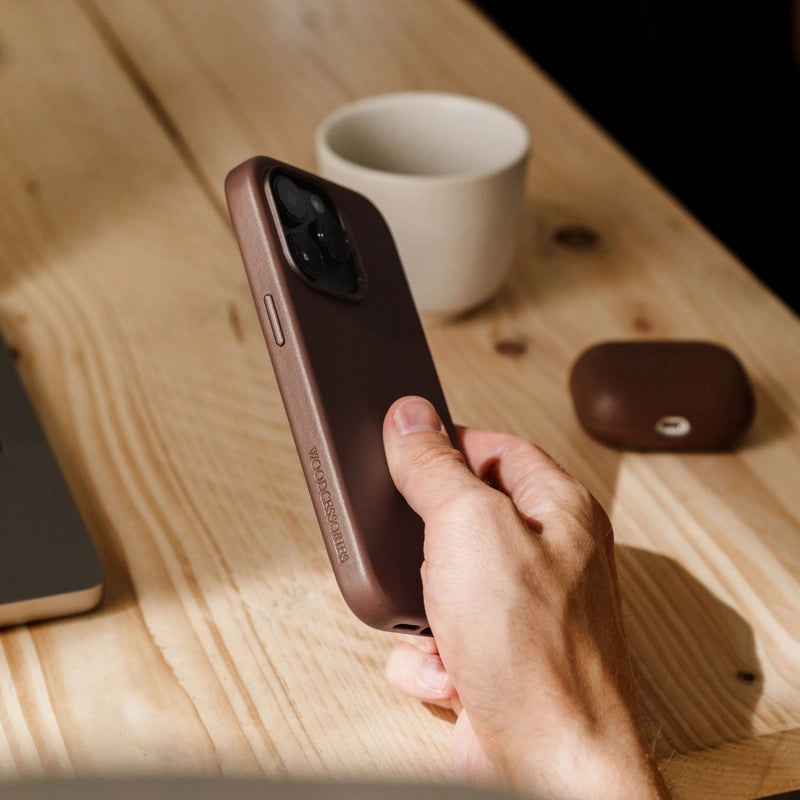 A person is holding an iPhone 17 Pro Max in a brown leather case. On the wooden table in the background are a cup and a brown leather AirPods case.