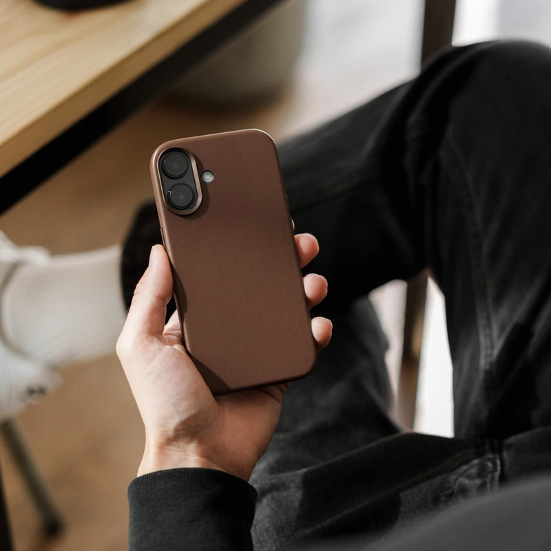 A person is holding an iPhone 16 in a brown leather case. The back of the phone shows the matte leather texture. In the out-of-focus background, you can make out a pair of dark jeans, a table leg, and a wooden floor.