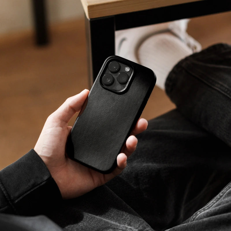 A person is holding an iPhone 16 Pro in a black leather case. A desk can be seen in the out-of-focus background.