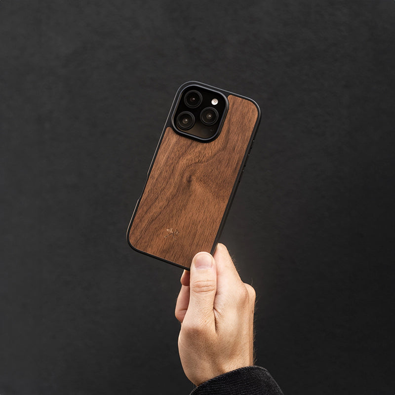 Gros plan sur une main tenant un iPhone 16 en finition « Bumper Case » avec du bois de noyer, sur fond sombre.