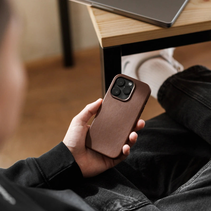Eine Person hält ein iPhone 16 Pro im braunen Leder 16 Pro Case in der Hand. Die Rückseite zeigt die matte Lederstruktur. Im unscharfen Hintergrund sind eine dunkle Jeans, ein Tischbein und ein Holzboden zu erkennen.