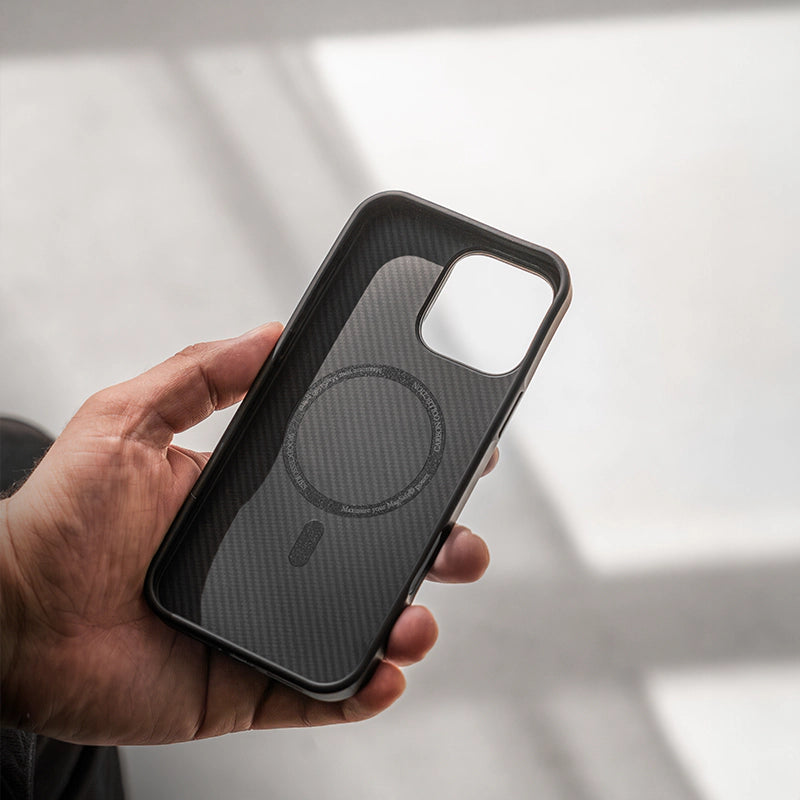 A hand is holding an empty, black iPhone 17 Air Carbon case and showing the inside, with the MagSafe ring clearly visible, against a bright, out-of-focus background.