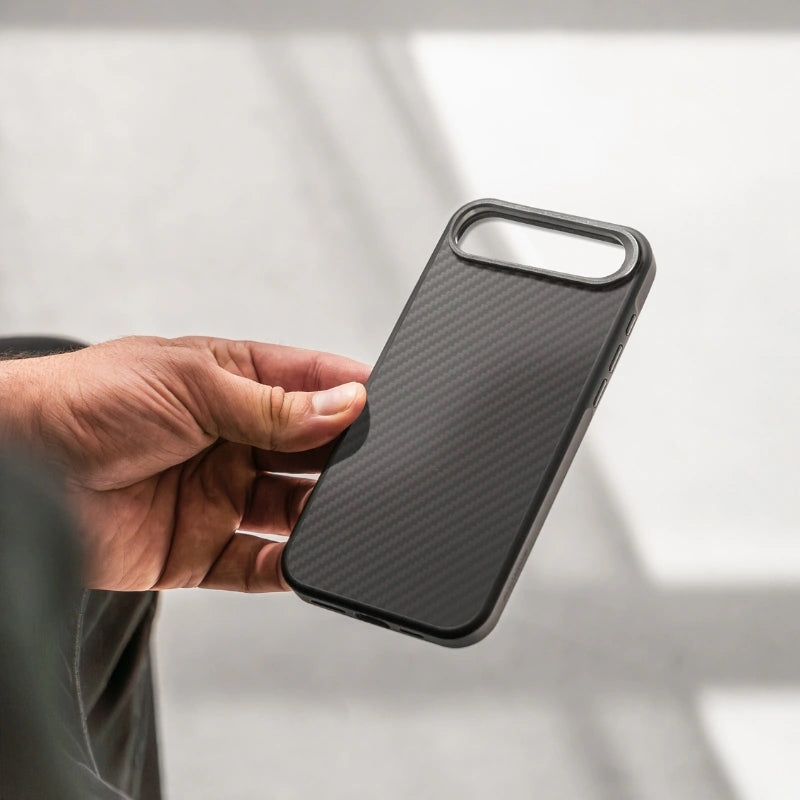 A hand is holding an empty, black carbon case for the iPhone 17 Air. The back shows the diagonal carbon weave pattern, while the wide, horizontal camera cutout at the top reveals a view of the bright, slightly blurred background.