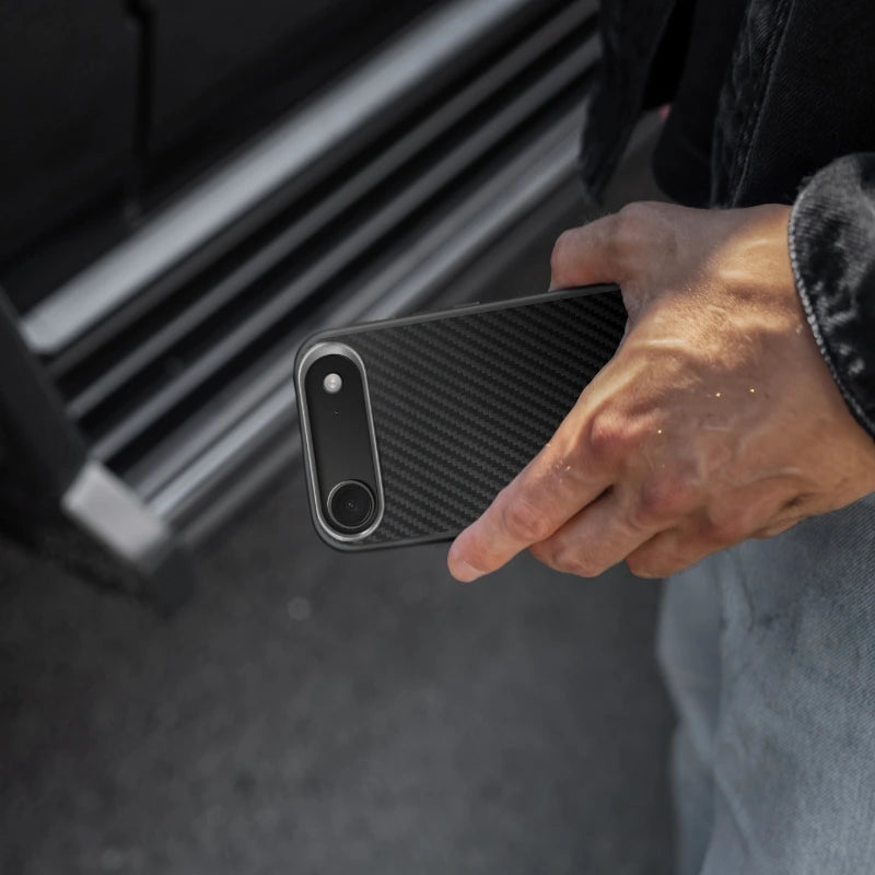 Une personne tient un iPhone 17 Air dans sa main, à l'horizontale, dans un étui noir en carbone. L'arrière du téléphone révèle la structure tissée caractéristique du carbone et l'encoche allongée abritant l'appareil photo. En arrière-plan, flou, on distingue le marchepied latéral d'une voiture.