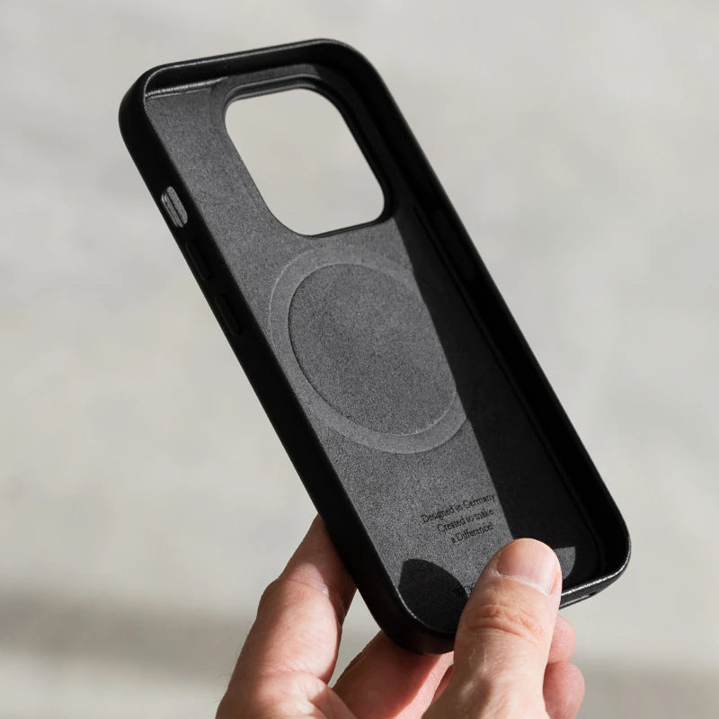 Eine Hand hält ein leeres, schwarzes iPhone 17 Leder-Case. Die Innenseite aus Filz zeigt einen MagSafe-Ring und einen kleinen Textaufdruck im unteren Bereich. Im unscharfen Hintergrund sind helle Steinstufen erkennbar.
