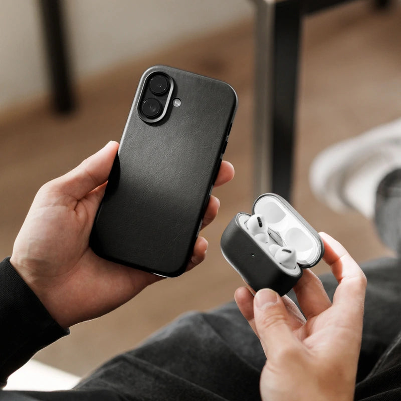 Zwei Hände halten ein schwarzes iPhone 17 im schwarzen Leder-Case sowie ein geöffnetes AirPods-Case, das ebenfalls in einer schwarzen Lederhülle steckt. Im unscharfen Hintergrund sind eine dunkle Hose und ein Holzboden zu sehen.