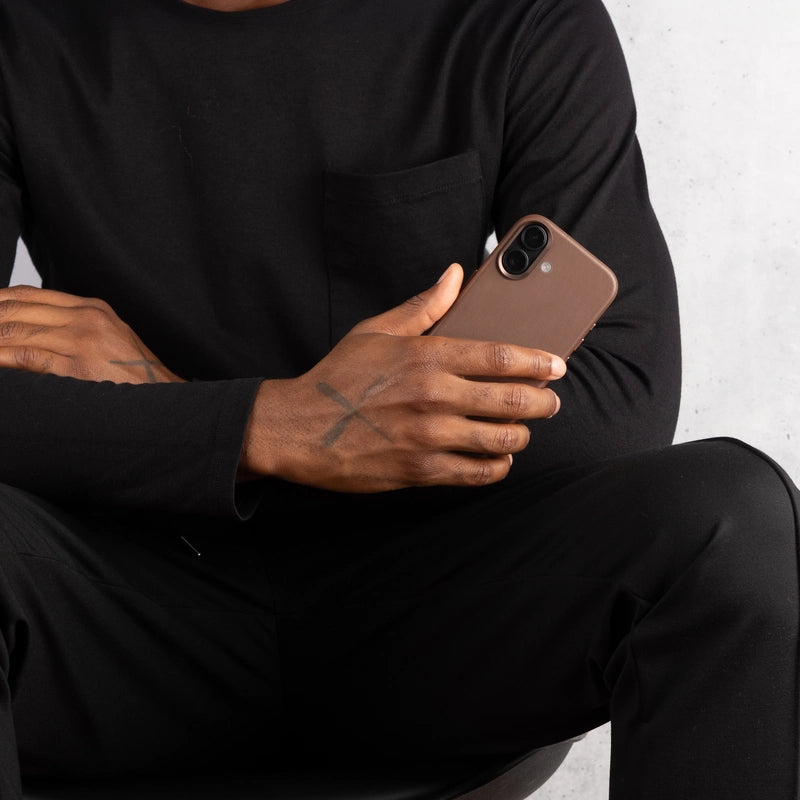 Eine in Schwarz gekleidete Person sitzt vor einem hellen Hintergrund und hält ein iPhone 17 Pro im braunen Leder-Case. Die braune Hüllenrückseite und der Kamera-Ausschnitt sind deutlich sichtbar.