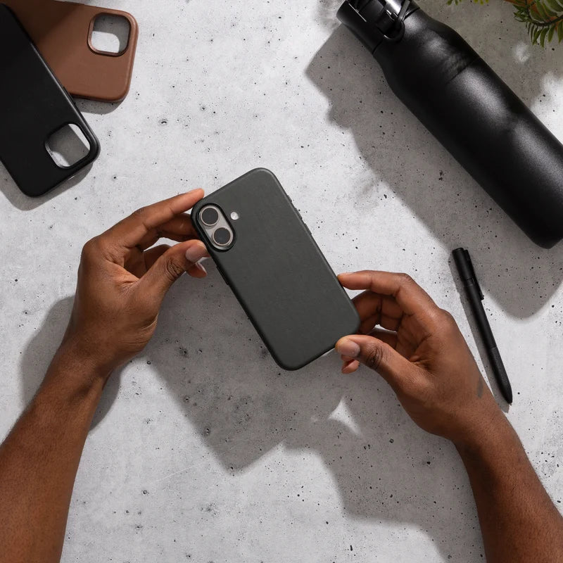 Zwei Hände halten ein schwarzes iPhone 17 im schwarzen Leder-Case über einer grauen Tischplatte in Betonoptik. Auf dem Tisch befinden sich außerdem eine schwarze Trinkflasche, ein schwarzer Stift sowie zwei weitere leere Cases in Schwarz und Braun.
