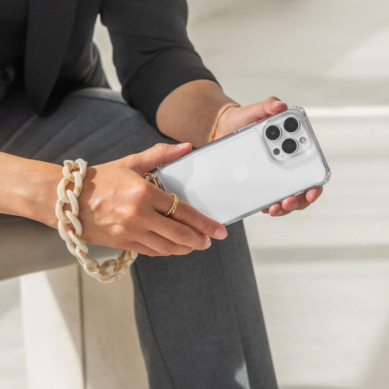 A person is holding a white iPhone 17 Pro horizontally in their hands, encased in the transparent “ Clear Case .” The short, chunky “ Crossbody Case ” made of resin with a marbled taupe finish wraps elegantly around the left wrist like a bracelet.