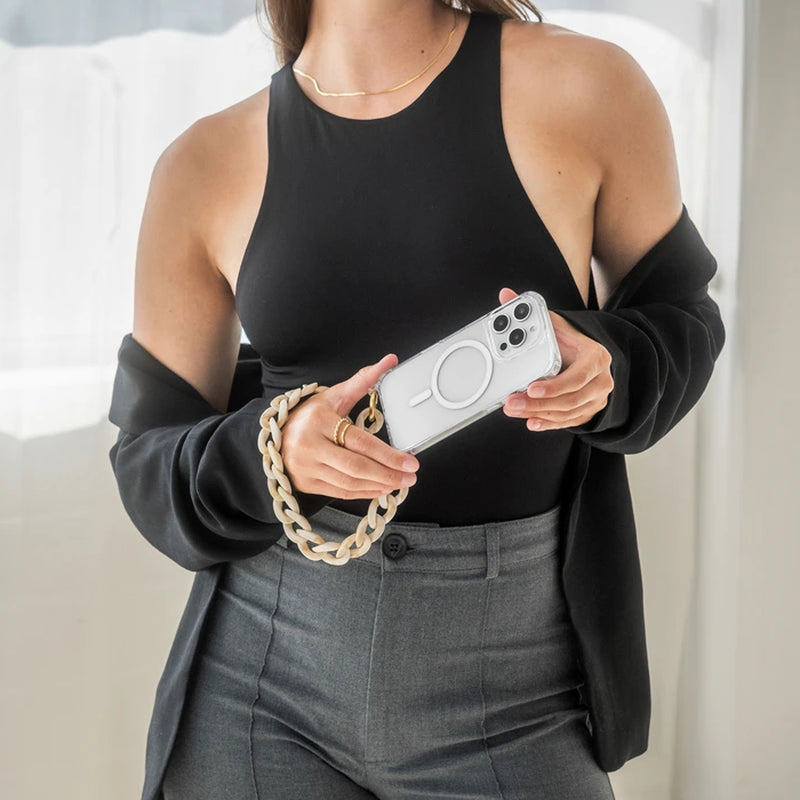 A person wearing a black top and gray pants is holding a white iPhone 17 Pro in a transparent Clear Case case, positioned diagonally in front of their body with both hands. The short, chunky Crossbody Case resin bracelet, with a marbled taupe finish, sits loosely around their right wrist like a thick bracelet.