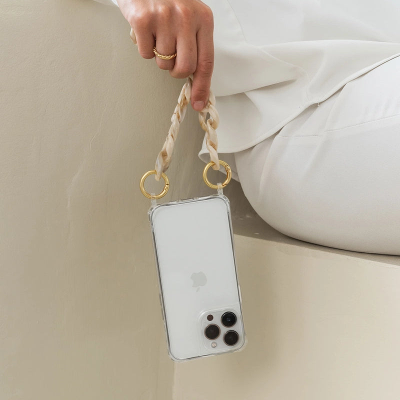A person wearing white clothing casually holds a white iPhone 17 Pro in a transparent Clear Case by their short, chunky Crossbody Case made of resin with a marbled taupe finish, so that the smartphone hangs upside down. Light, minimalist background.