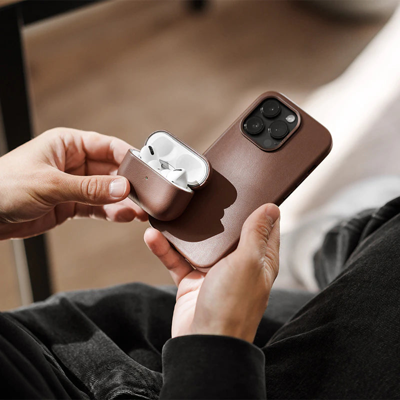 Two hands are holding a black iPhone 17 Pro in a brown leather case, as well as an open AirPods case, which is also inside a brown leather sleeve. In the out-of-focus background, a pair of dark pants and a wooden floor are visible.