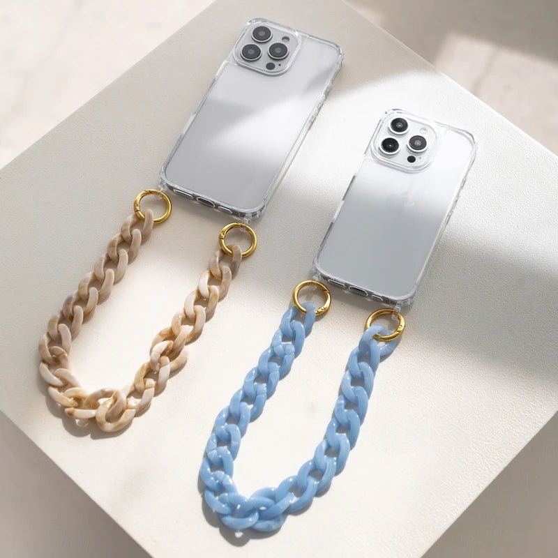 Two white iPhone 17 Pro Max phones in transparent clear cases lie side by side on a light-colored surface, with their backs facing up. Attached to the left case is a short, chunky “ Crossbody Case ” made of resin with a marbled taupe finish; attached to the right case is an identical chain in light blue. Both are attached to the “ Case ” with gold rings.