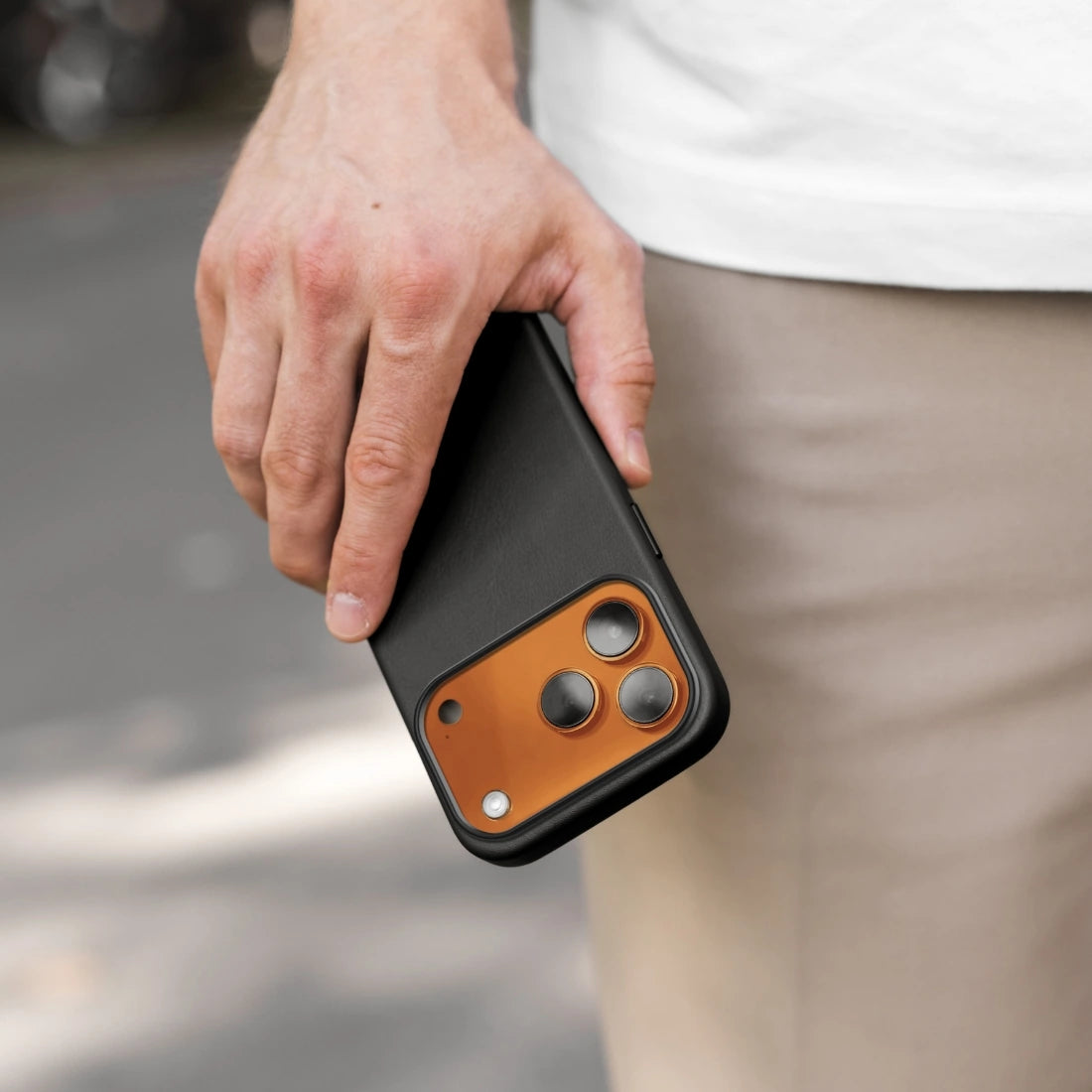 A hand is holding an orange iPhone 17 Pro in a black leather case outdoors. The orange back of the device is visible through the large camera cutout. In the out-of-focus background, light-colored pants and pavement can be seen.