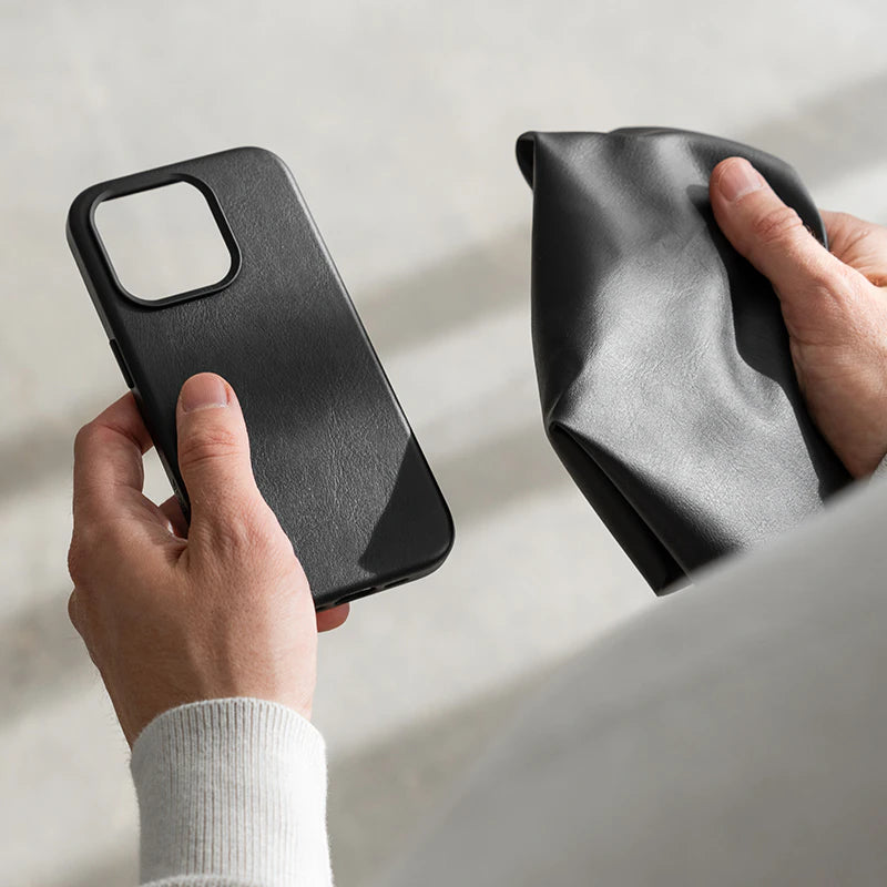 Eine Hand hält ein leeres, schwarzes Leder-Case für das iPhone 17. Eine zweite Hand daneben hält ein Stück schwarzes Leder. Im Hintergrund sind unscharfe, helle Steinstufen zu erkennen.